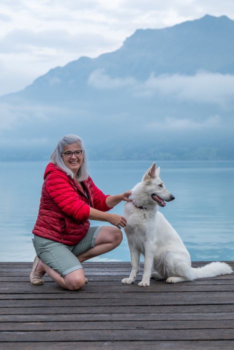 Silvia in Roter Jacke schau nach vorn. Rechts neben ihr Jira (Schäferhund) schaut in die Ferne. im Hintergrund ein idyllischer See und Berge